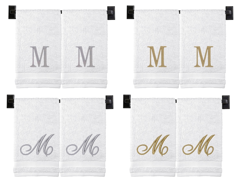 May include: Set of four white hand towels with embroidered letter "M" in silver and gold. Two towels feature a simple "M" and two towels feature a cursive "M".