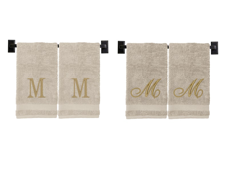 May include: Two beige hand towels with embroidered gold "M" initials. One towel has a block letter "M" and the other has a cursive "M".