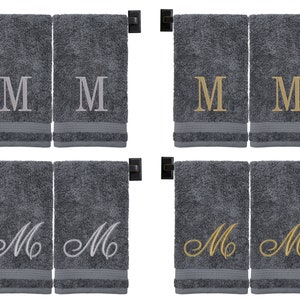 May include: Four gray hand towels with embroidered letter "M" in different styles. Two towels have a simple block letter "M" in silver, two have a script "M" in gold, and two have a script "M" in silver.