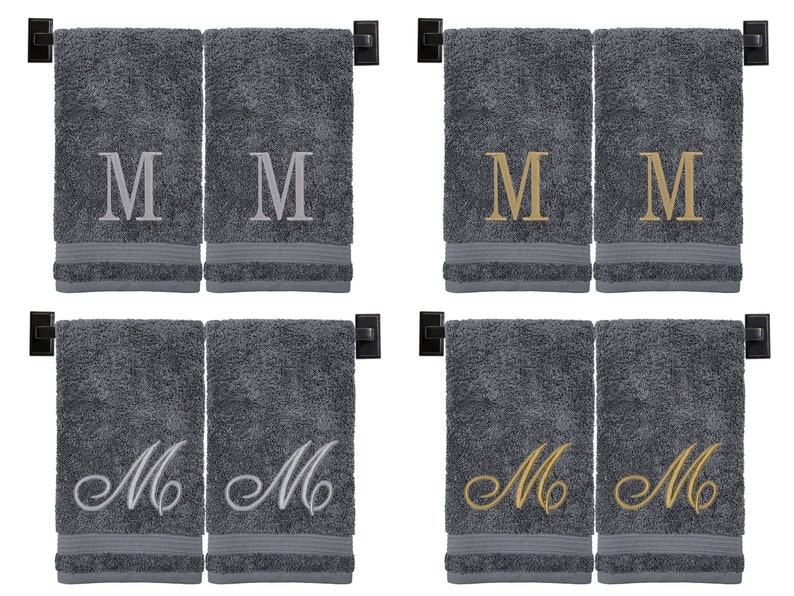 May include: Four gray hand towels with embroidered letter "M" in different styles. Two towels have a simple block letter "M" in silver, two have a script "M" in gold, and two have a script "M" in silver.