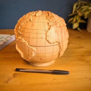 Puede incluir: Un globo terráqueo esférico de madera con continentes tallados en relieve y líneas de cuadrícula grabadas. La madera de tono claro muestra vetas naturales. Un bolígrafo negro está colocado horizontalmente frente al objeto.