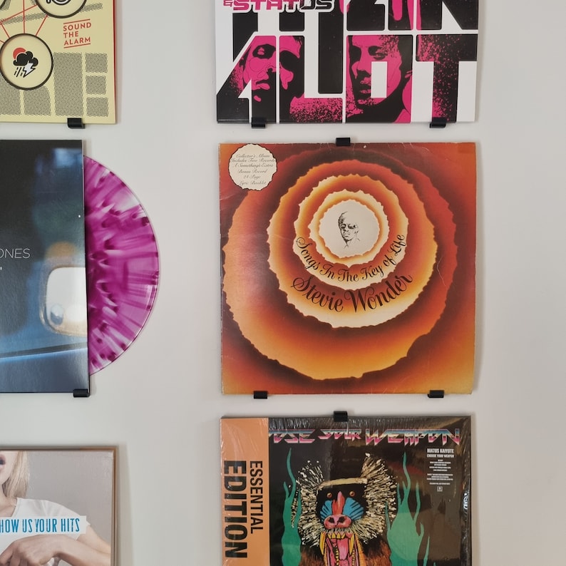 May include: A collection of vinyl records displayed on a wall using black metal record holders. The records include "Status Quo - In the Army Now", "Stevie Wonder - Songs in the Key of Life", and "Essential Edition - The Four Tops".