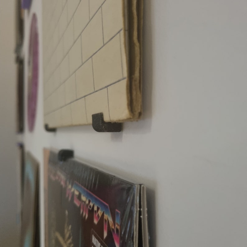 May include: Close-up of a black vinyl record holder mounted on a white wall. The holder is made of plastic and has a simple design. The record is a black vinyl record with a white label.