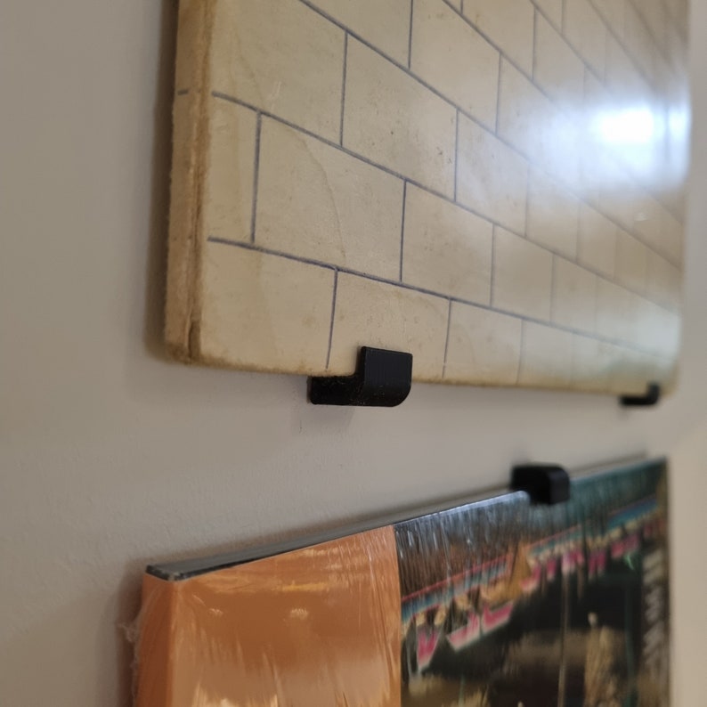 May include: Two vinyl records are displayed on a wall using black plastic record holders. The top record has a white brick pattern, and the bottom record has a colorful design.