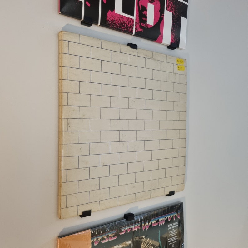 May include: A white brick wall record album cover with black trim. The album cover is hanging on a wall with other record album covers.