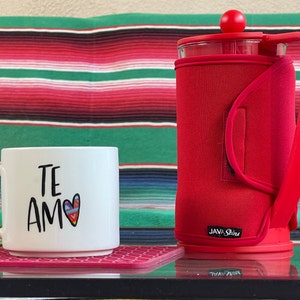 May include: A red insulated coffee press with a black handle and the text "Java Skins" on the bottom. A white mug with the text "Te Amo" and a colorful heart design is on a pink coaster.