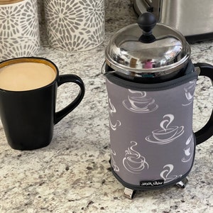 May include: A gray and white insulated coffee press with a black handle and a silver lid. The press has a pattern of white coffee cups with steam coming from them. The text "JAVA Shine" is printed on the bottom of the press.