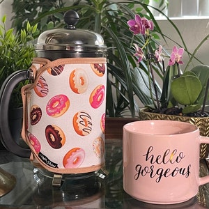 May include: A stainless steel French press coffee maker with a donut-patterned fabric sleeve. The sleeve has pink, brown, and white donuts on a white background. The text "Java Street" is printed on the sleeve. The coffee maker is sitting on a table next to a pink ceramic mug with the text "hello gorgeous" printed on it.