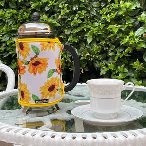 May include: A white ceramic coffee pot with a black handle and a yellow and white sunflower print. The pot is sitting on a white table with a glass top. The text "JAVA Shine" is printed on the pot.