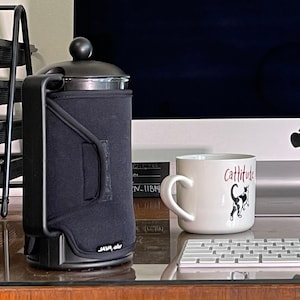May include: A black French press coffee maker with a black insulated sleeve, next to a white mug. The mug has the word "Cattitude" and a black and white cat illustration. A keyboard and computer are in the background.