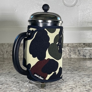 May include: A stainless steel French press coffee maker with a black and green camouflage fabric sleeve. The sleeve has the text "JAVA Skins" printed on it.