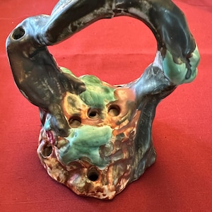 May include: A ceramic flower frog with a dark brown handle and a base with multiple holes. The base features green, brown, and red accents. The handle is arched, creating a circular opening.