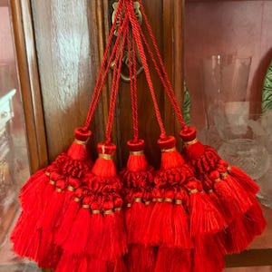 以下が含まれることがあります： 装飾的な真鍮製の金具から吊り下げられた、金色のアクセントが付いた6つの鮮やかな赤いタッセル。タッセルは、層状のテクスチャード加工された生地で作られており、ふっくらとした華やかな外観を作り出しています。背景は木製のキャビネットです。