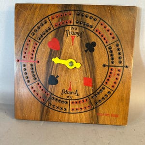 Könnte beinhalten: Ein Vintage-Cribbage-Brett aus Holz mit einer kreisförmigen Bahn und Lochungen für die Stifte. Das Brett zeigt Kartensymbole, die Worte "No Trumps" und "Spades On" sowie einen gelben Stifthalter. Das Brett ist quadratisch und hat eine Holzmaserung.
