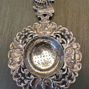 May include: Ornate silver tea strainer with a ship motif. The strainer features a circular bowl with small holes, surrounded by an elaborate floral and scroll design. A detailed ship tops the handle, adding to its decorative appeal.