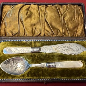 May include: An antique silver serving set, including a knife and a spoon, presented in a velvet-lined box. The knife and spoon have mother-of-pearl handles and ornate floral designs. The box is dark with a gold interior.