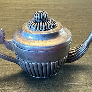 May include: A miniature silver teapot with intricate detailing. It has a curved spout, a decorative handle, and a lid with a textured knob. The body of the teapot is adorned with vertical grooves. A charming decorative item.