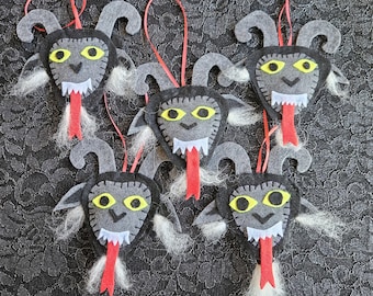 May include: Six gray felt ornaments shaped like a goat's head with yellow eyes, white teeth, and a red tongue. Each ornament has a red ribbon attached.