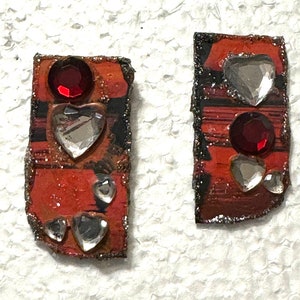 May include: A pair of red and black rectangular earrings with silver glitter. Each earring has a red rhinestone, a clear heart-shaped rhinestone, and two smaller clear rhinestones.