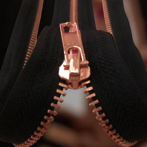 Puede incluir: Primer plano de una cremallera de oro rosa con tela negra a cada lado. La cremallera está abierta y la tela está doblada hacia atrás.