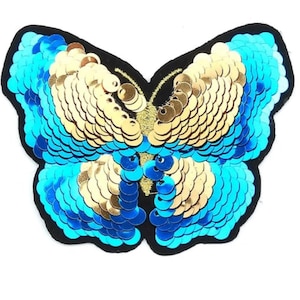 Può includere: Un cerotto a forma di farfalla con paillettes blu e dorate. La farfalla è bordata di nero e ha uno sfondo nero.
