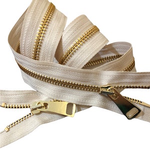 Puede incluir: Primer plano de una cremallera beige con dientes de metal dorado. La cremallera está enrollada y el deslizador es visible.