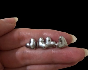 May include: Four silver heart-shaped earrings, displayed on a hand.