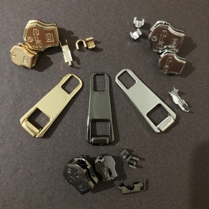 Può includere: Assortimento di tiretti per cerniere lampo in metallo dorato, argentato e nero. I tiretti sono di varie forme e dimensioni, tra cui uno slider, un fermo superiore e un fermo inferiore.