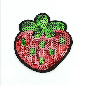 Può includere: Un toppa a forma di fragola rossa e verde con paillettes. La fragola è bordata di nero e ha una foglia verde con paillettes in cima.