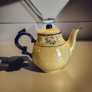 Puede incluir: Una tetera de cerámica amarilla con asa y tapa azules. La tetera tiene un diseño floral y un ribete azul oscuro. Un diseño de tetera clásico, ideal para servir té.