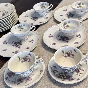 May include: A vintage porcelain tea set featuring a floral pattern in shades of purple and green. The set includes teacups, saucers, plates, and a gravy boat. The teacups have gold trim. The plates are oval-shaped.