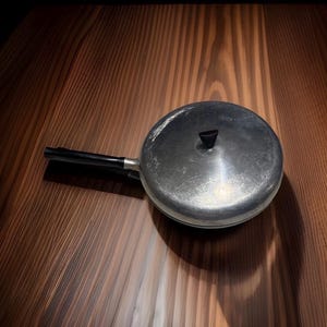 May include: A round, silver-colored metal pan with a lid and a long, black handle. The lid features a small, black, triangular-shaped knob in the center. The surface of the metal pan shows signs of wear and light scratches across the top.