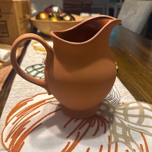 May include: A terracotta-colored pitcher with a curved handle and a spout. The pitcher sits on a table runner with an embroidered pumpkin design in orange and white. The pitcher is a warm, earthy tone.