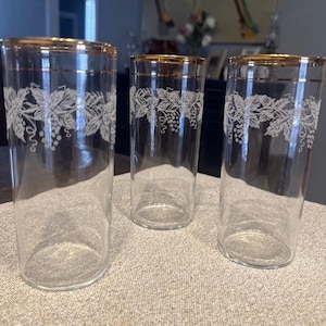 May include: Three clear glass tumblers featuring a white etched grape vine pattern around the upper portion and a thin gold rim at the top edge. The glasses are cylindrical in shape with smooth walls and a flat base, arranged in a group of three.