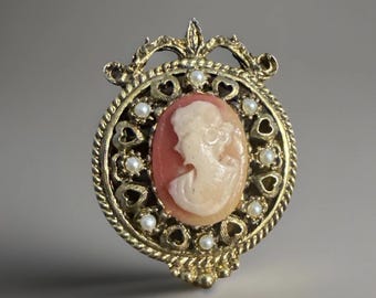 Puede incluir: Broche de camafeo dorado vintage. El camafeo ovalado presenta el perfil de una mujer en crema sobre un fondo salmón. El broche está enmarcado por un diseño en forma de corazón, pequeñas perlas y un borde de cuerda trenzada. Una parte superior decorativa completa el diseño.