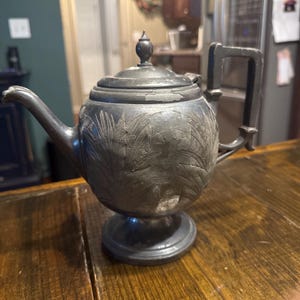 May include: A dark gray metal teapot featuring a rounded body with an embossed botanical leaf pattern, a pedestal base, a curved spout, and a lid with a finial. The surface shows signs of wear and paint loss, revealing a lighter metallic finish.
