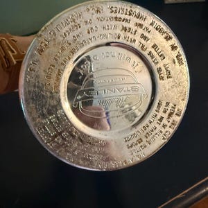 Puede incluir: La base circular de color plateado de una botella t&eacute;rmica Stanley Aladdin. La superficie met&aacute;lica presenta texto en relieve que incluye 'Stanley', 'Aladdin', 'No. A-943A Pint' e instrucciones de uso como 'It will not break' y 'Nashville, Tenn. U.S.A.'