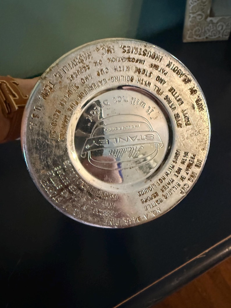 Puede incluir: La base circular de color plateado de una botella t&eacute;rmica Stanley Aladdin. La superficie met&aacute;lica presenta texto en relieve que incluye 'Stanley', 'Aladdin', 'No. A-943A Pint' e instrucciones de uso como 'It will not break' y 'Nashville, Tenn. U.S.A.'