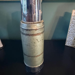 Puede incluir: Un termo cil&iacute;ndrico vintage con un cuerpo de metal verde claro desgastado que presenta bandas horizontales de &oacute;xido y desgaste. El recipiente est&aacute; rematado con una tapa de cromo pulido y reflectante y tiene una base de cromo a juego.