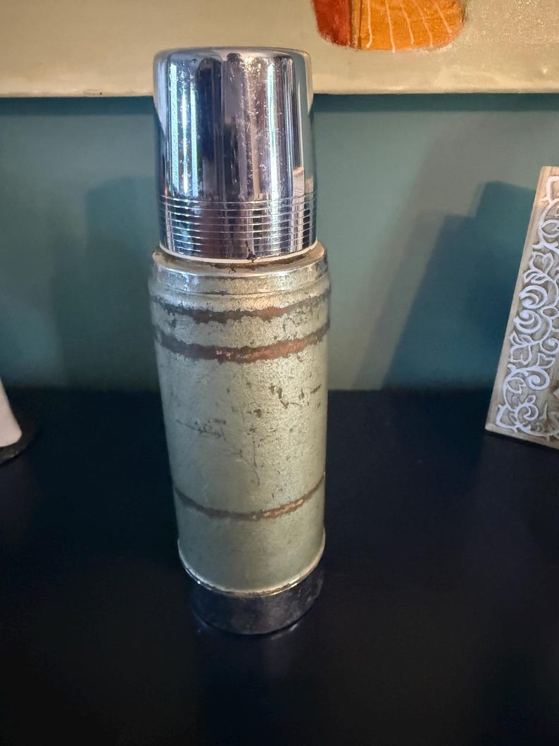 Puede incluir: Un termo cil&iacute;ndrico vintage con un cuerpo de metal verde claro desgastado que presenta bandas horizontales de &oacute;xido y desgaste. El recipiente est&aacute; rematado con una tapa de cromo pulido y reflectante y tiene una base de cromo a juego.