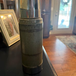 Puede incluir: Un termo cil&iacute;ndrico vintage con un cuerpo de metal verde apagado y desgastado, y una tapa de cromo pulido. El exterior muestra signos de desgaste con textura visible y bandas horizontales. La base presenta un borde de cromo a juego.