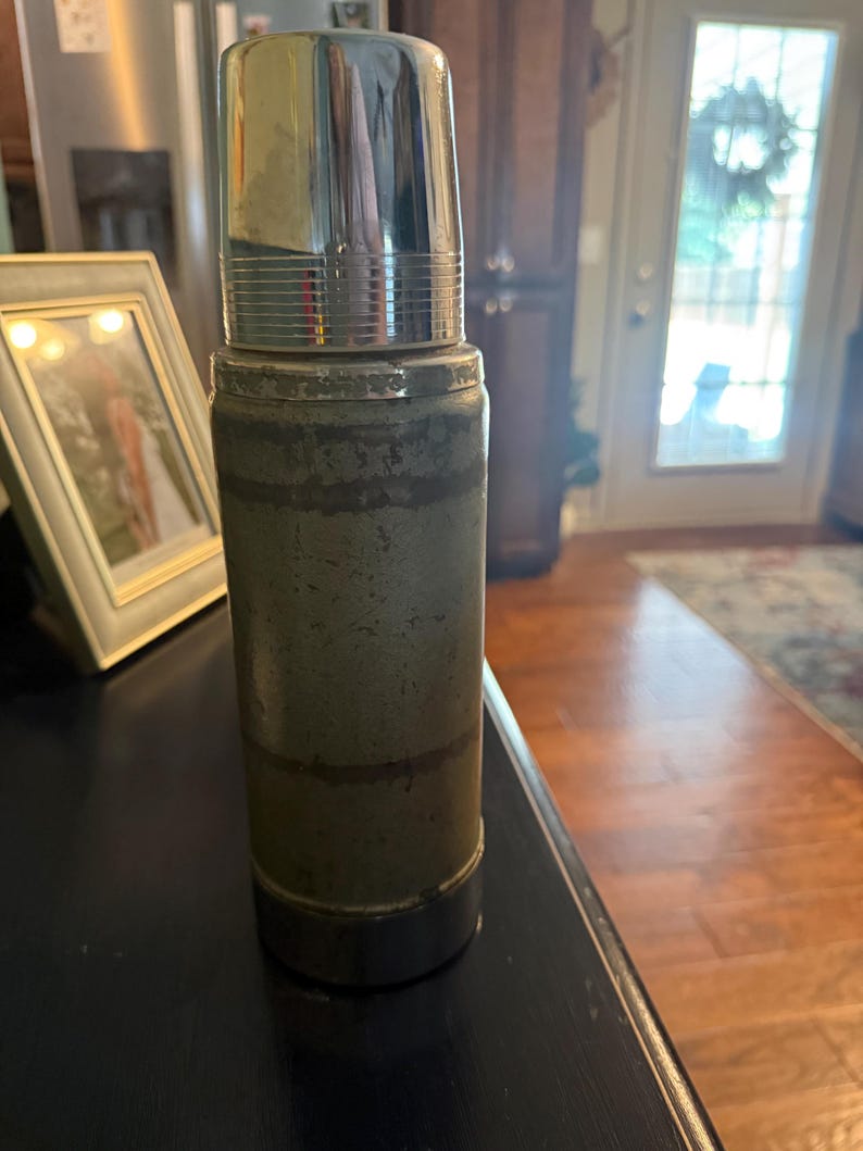 Puede incluir: Un termo cil&iacute;ndrico vintage con un cuerpo de metal verde apagado y desgastado, y una tapa de cromo pulido. El exterior muestra signos de desgaste con textura visible y bandas horizontales. La base presenta un borde de cromo a juego.