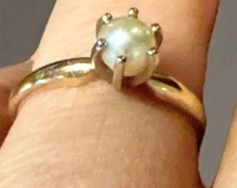 May include: A gold ring featuring a round, off-white pearl. The pearl is held in place by six prongs. The ring's band is a simple, polished gold. This piece of jewelry is a classic and elegant design.