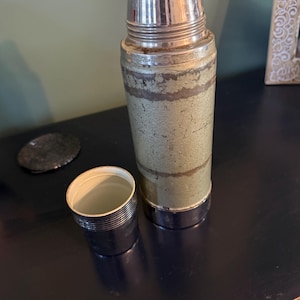 Puede incluir: Un termo vintage con cuerpo de metal texturizado en color verde salvia que presenta desgaste y ara&ntilde;azos visibles. Tiene el cuello y la base de cromo pulido, rematado con una tapa de pl&aacute;stico color crema. Una taza cromada a juego se sit&uacute;a al lado.