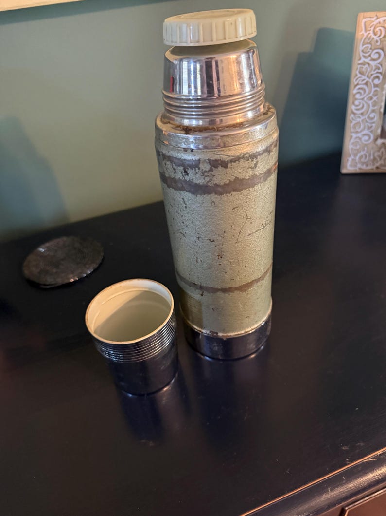 Puede incluir: Un termo vintage con cuerpo de metal texturizado en color verde salvia que presenta desgaste y ara&ntilde;azos visibles. Tiene el cuello y la base de cromo pulido, rematado con una tapa de pl&aacute;stico color crema. Una taza cromada a juego se sit&uacute;a al lado.