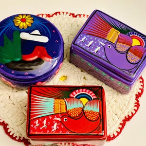 May include: Three hand-painted wooden boxes with colorful designs. The boxes are decorated with birds, flowers, and geometric patterns. The boxes are a vibrant purple, red, and blue.
