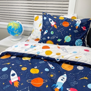 May include: A blue and white space-themed duvet cover with a navy blue background and white stars. The duvet cover features rockets, planets, and stars. The duvet cover is shown on a bed with a white and blue pillowcase with a space-themed design.