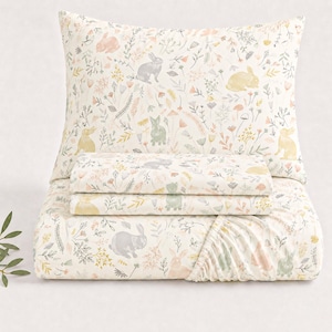 Op de afbeelding: Een crèmekleurige beddengoedset met een bloemen- en konijnenpatroon. De set bevat een kussensloop, een hoeslaken, een laken en een dekbedovertrek. Het ontwerp bevat pastelkleuren zoals roze, geel en grijs.
