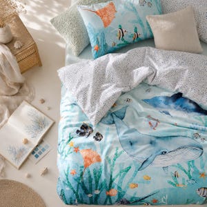 Puede incluir: Un juego de funda nórdica y almohadas azul claro con temática submarina. La ropa de cama presenta ilustraciones de ballenas, delfines, rayas y varios peces. Las almohadas son en tonos verdes y beige. También se ve un libro con ilustraciones de coral.