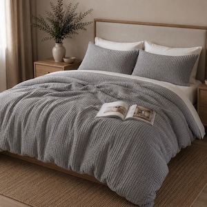 May include: A bed dressed with a grey waffle-weave duvet cover and matching pillowcases. White sheets are visible beneath the duvet. An open book rests on the bed. The bedroom has neutral tones and wooden bedside tables.